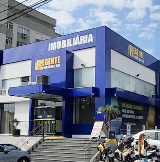 "Fachada da imobiliária Regente Imóveis, com uma construção moderna de cor azul e amarela, localizada em uma esquina com degraus de acesso e uma placa de 'vende-se' na entrada."